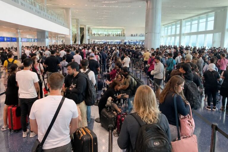 Crowded airport terminal with long lines of travelers and rolling suitcases awaiting check-in or security.