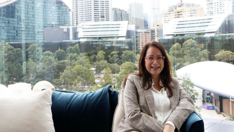 Woman with glasses seated on a blue sofa indoors, smiling, with a city park and high-rise buildings visible through a glass wall behind her.