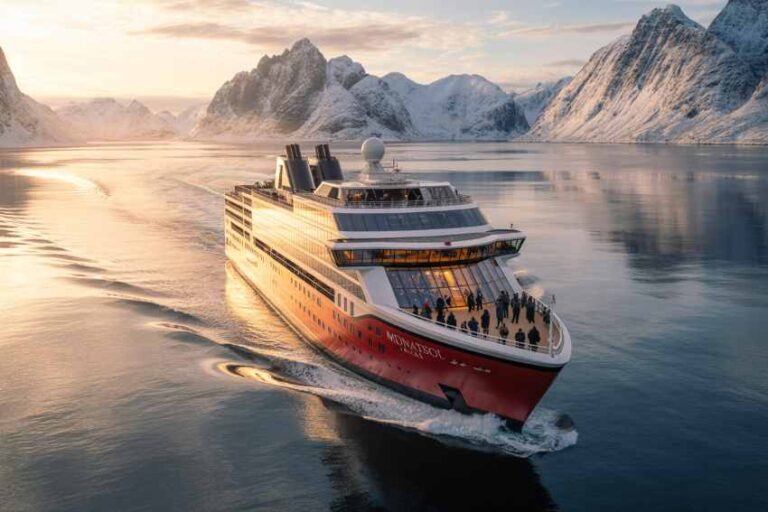 Large red-and-white cruise ship sailing through a calm fjord with snow-covered mountains at sunset; passengers on deck.