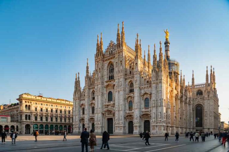 Milano Cathedral (Duomo di Milano) facade bathed in sunlight with pilgrims in Piazza del Duomo, Milan, Italy to the left and people walking around the square.