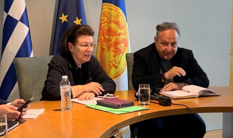 Two people sit at a curved conference table during a meeting, with notebooks, a water bottle, and flags in the background.