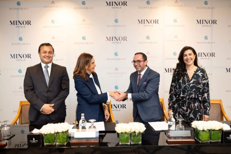 Business signing ceremony: two suited professionals shake hands across a table while two others stand smiling behind them, backdrop with hotel logos A–Z.