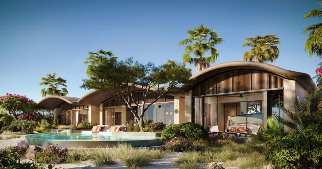 Luxurious beachfront villa with curved roofs, glass walls, and a shallow pool surrounded by tropical landscaping under a clear sky