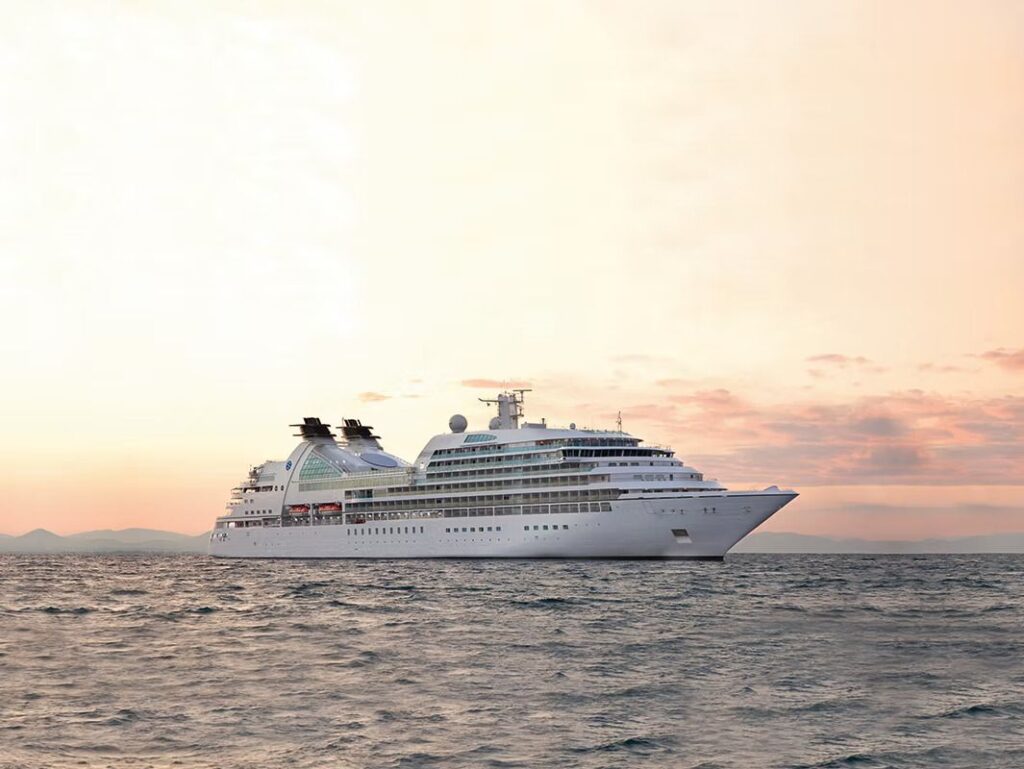 Large white cruise ship sailing on calm sea at sunset with pink and orange sky in the background.