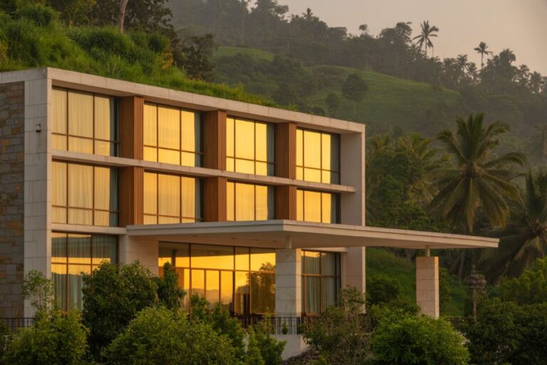 Modern two-story house with large glass windows reflecting a golden sunset, set among green hills and palm trees.