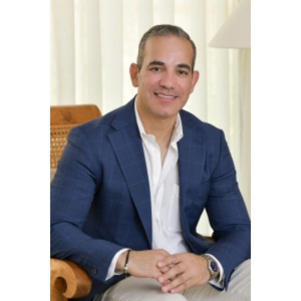Professional headshot: smiling man in a navy blazer and white shirt, seated indoors with light curtains behind him, hands folded.