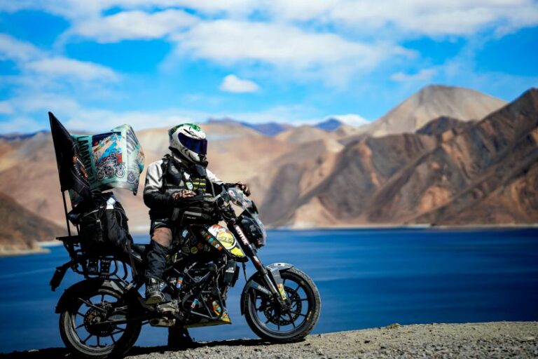 Motorcyclist in full riding gear on a loaded adventure bike, parked by a blue lake with rugged mountains in the background.