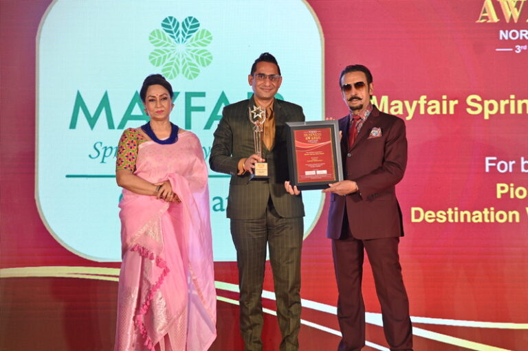 Three people on a stage during an awards ceremony: a man in a green suit holds a trophy while another man in a maroon suit presents a plaque; a woman in a pink sari looks on.