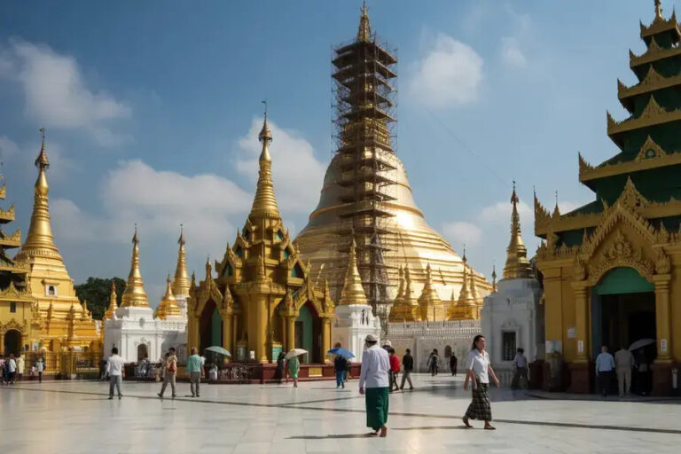 Golden Buddhist temple courtyard with tall gilded stupas, scaffolding on the central dome, and visitors walking on white marble.