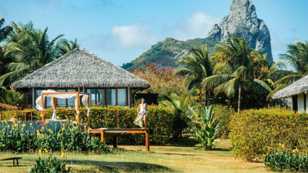 Tropical resort with thatched-roof bungalows, lush plants, and a person walking along a wooden deck.
