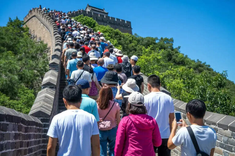 Crowd of tourists walking along a stone section of the Great Wall of China on a sunny day, with blue sky above.