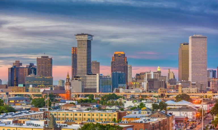 City skyline at dusk with tall skyscrapers, orange-pink sky, and low-rise foreground buildings.