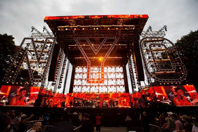 Large outdoor concert stage with red lighting, illuminated geometric logos, and a crowd in front of the stage at dusk/evening.