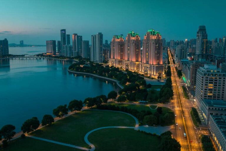 Evening cityscape along a river with illuminated skyscrapers and a park in the foreground.