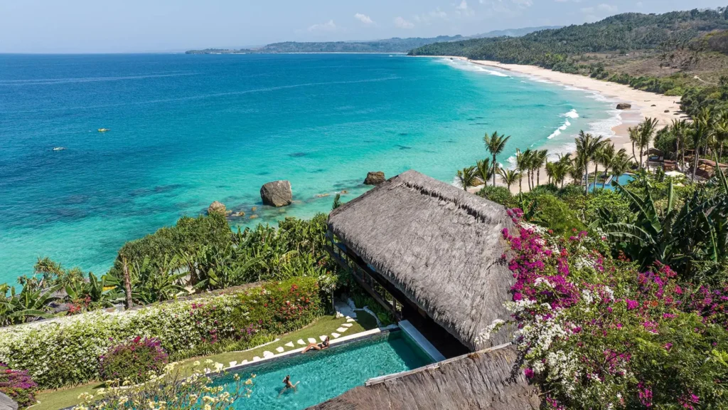 Bird's-eye view of a tropical resort perched above a turquoise sea, with a thatched-roof building, a swimming pool, and vibrant flowering plants along the garden.