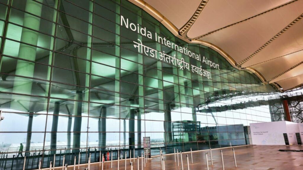 Noida International Airport sign on a glass façade with green-tinted pillars and a curved beige roof overhead (airport terminal exterior).