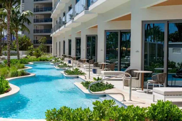 Resort pool winding along a modern white building with balconies, palm trees and wicker seating nearby.
