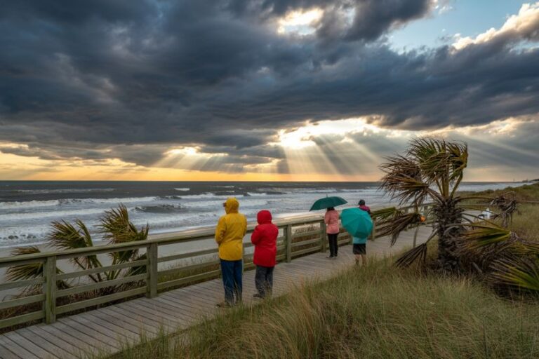 People in bright rain jackets stand on a wooden boardwalk watching rough seas as sun rays break through dark clouds.