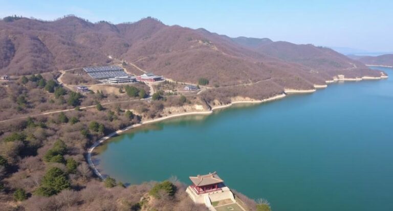 Aerial view of a blue lake edged by brown hills; a small red-roofed pavilion sits at the water's edge with buildings on the hillside nearby.