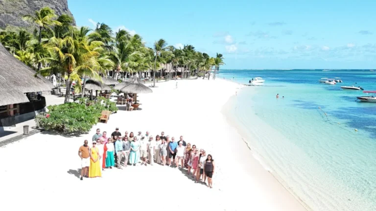 Group of people posing together on a sunny white-sand tropical beach with palm trees and thatched huts.