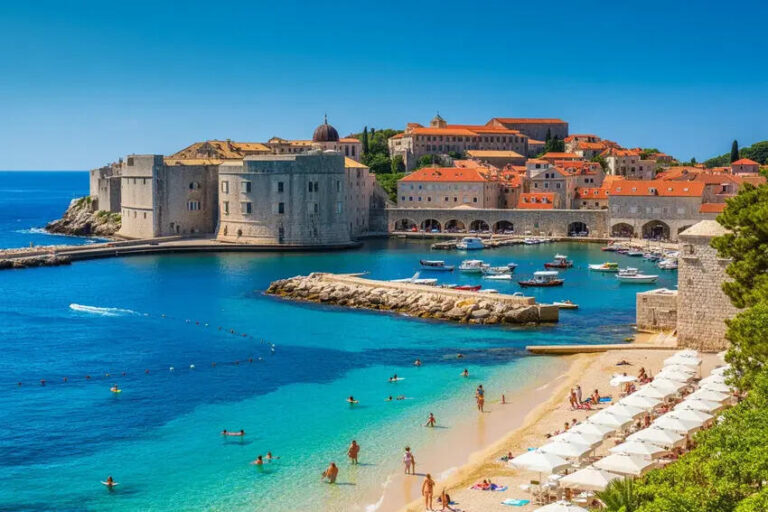 Historic fortress and old town by a blue-green harbor, with small boats, a sandy beach, and white umbrellas lining the shore.