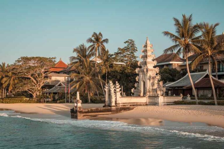 White stone pagoda shrine on a sandy beach, palm trees and calm turquoise sea in the background.