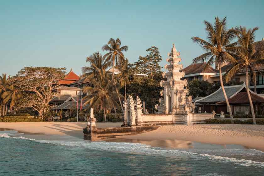 White stone pagoda shrine on a sandy beach, palm trees and calm turquoise sea in the background.