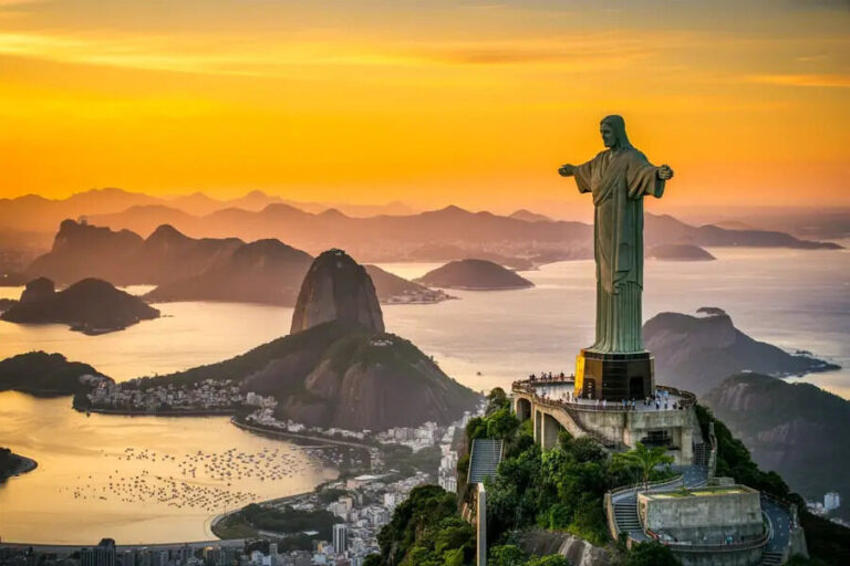 Christ the Redeemer statue with outstretched arms atop Corcovado Mountain overlooking Guanabara Bay at sunset.