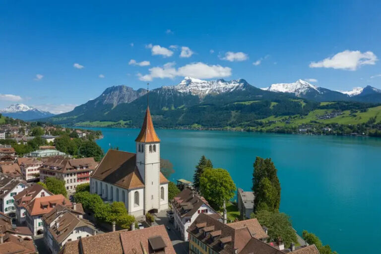 Aerial view of a lakeside town with a white church and red-tiled rooftops, turquoise lake, and snow-capped mountains in the distance under a blue sky.