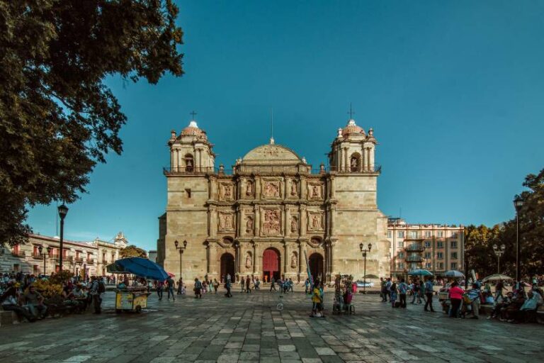 Front facade of a grand historic cathedral with red doors, in a busy plaza filled with people and stalls under a clear blue sky.