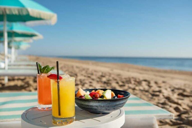 Two colorful tropical drinks with a bowl of fruit on a striped table at a sunny beach.