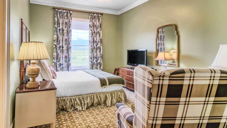 Hotel room with a queen bed, nightstands, lamps, dresser with a TV, floral curtains, and a plaid armchair/loveseat nearby.