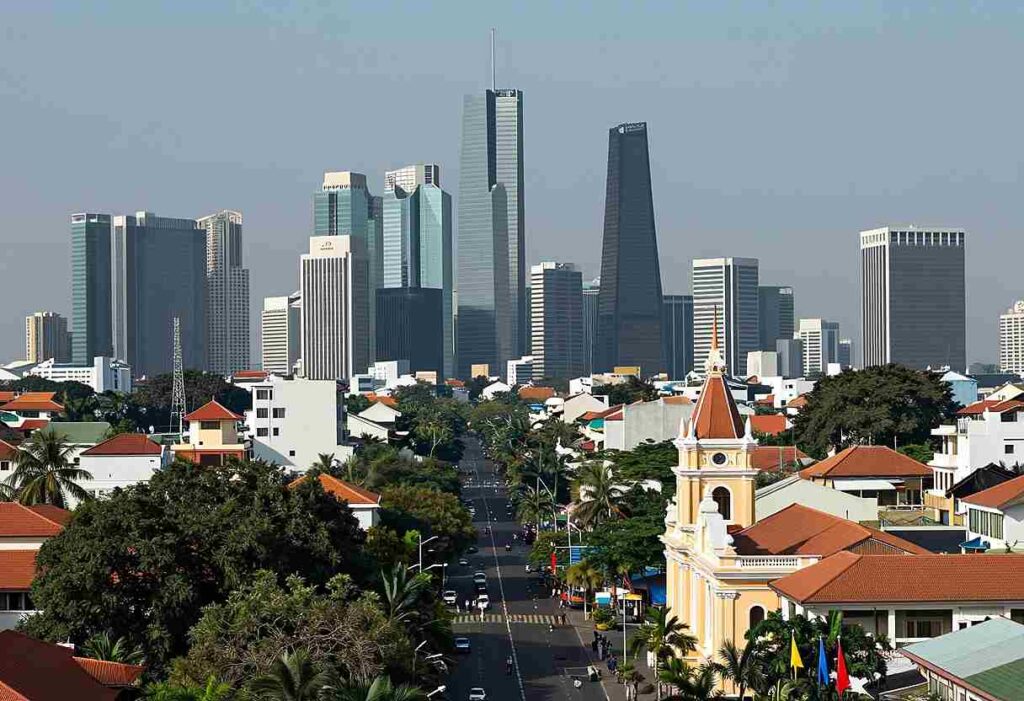 Aerial view of a downtown skyline with tall glass skyscrapers behind a neighborhood of red-tiled houses and a church with a clock tower in the foreground.