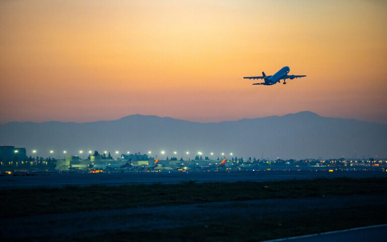 Airplane taking off from a sunset-lit airport with runway lights and distant hills in the background.