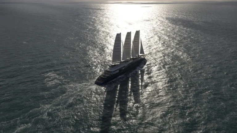 A large four-masted luxury sailing yacht gliding on the sea, sunlight reflecting off the water behind it.