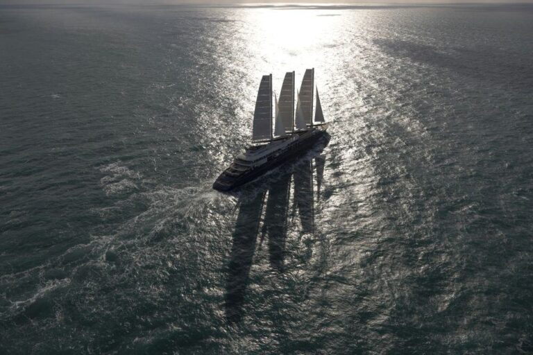 Four-masted sailing yacht cutting through sunlit ocean, casting long shadow on the water.