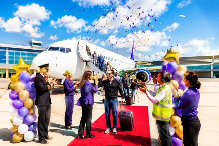 Celebration on the airport tarmac as travelers arrive by a white airplane, with a red carpet, balloons, and confetti.