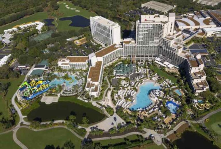 Aerial view of a large resort with multiple pools, water slides, and white hotel buildings surrounded by greenery and golf links nearby.