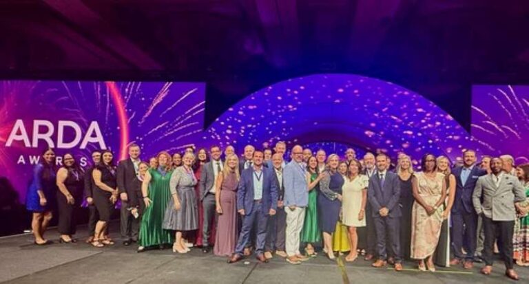 Group of formally dressed people on a stage posing for a photo at an ARDA Awards event with a purple backdrop and fireworks design