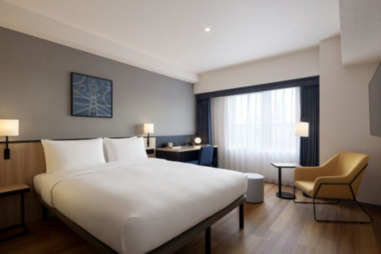 King-size bed with white bedding in a modern hotel room, gray accent wall, bedside lamps, and a desk area by the window.