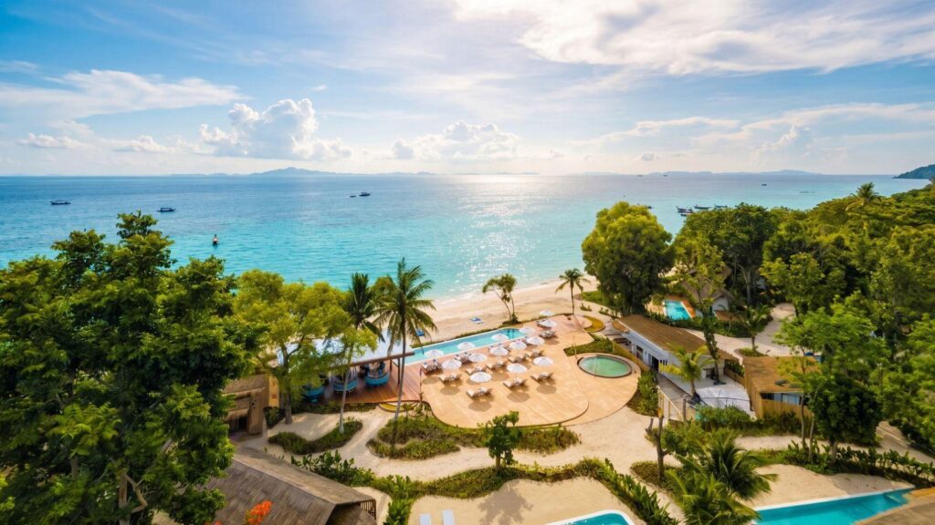 A tropical beach resort with turquoise ocean, white sand, palm trees, and a poolside area with umbrellas and sun loungers.