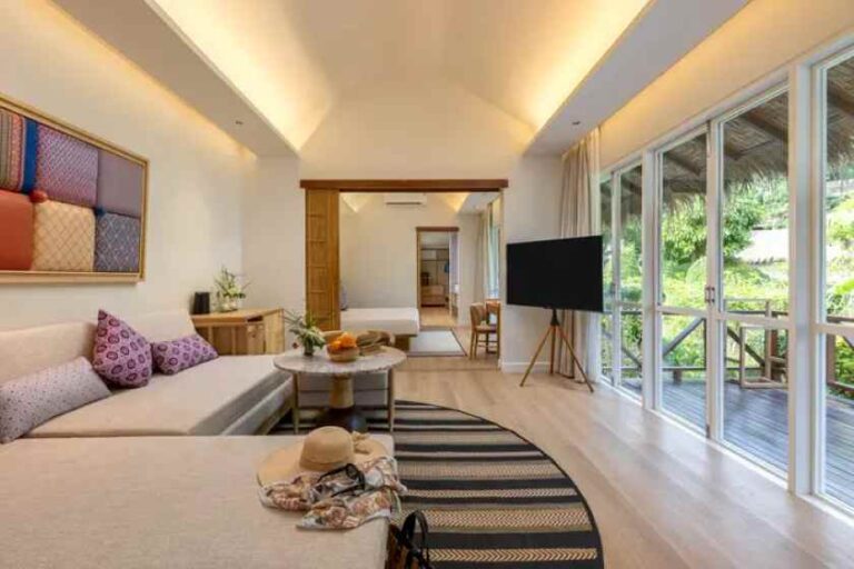 Open-plan hotel suite: beige sofa with purple cushions, round marble coffee table, and a striped rug facing a flat-screen TV on a stand near floor-to-ceiling windows.