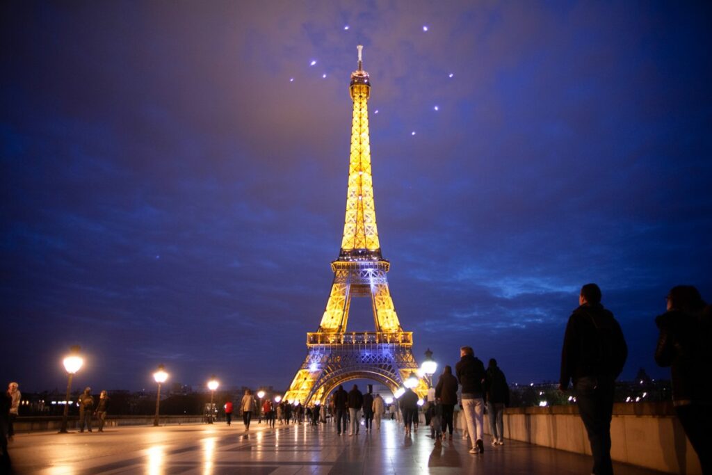 Eiffel Tower lit in warm yellow lights at night, with people walking along a waterfront promenade under a dark blue sky.