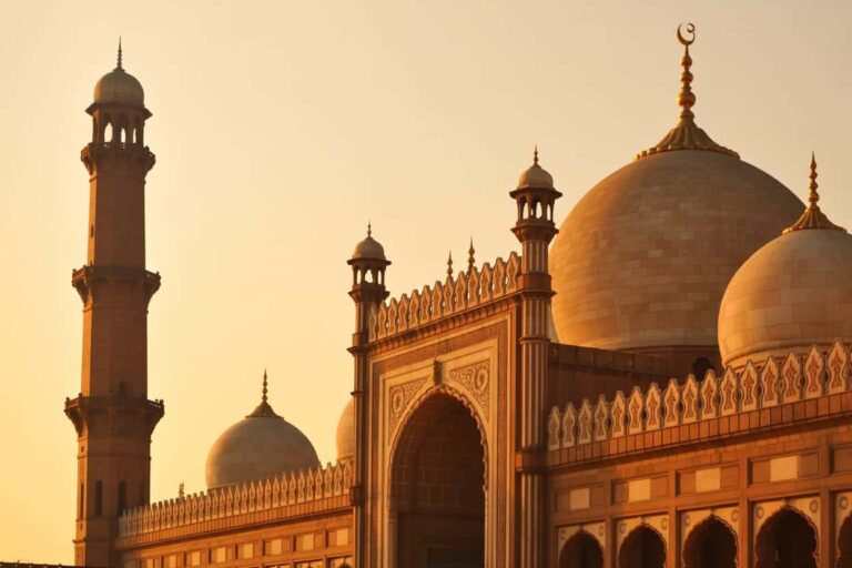 Sunset-lit mosque with a large central dome, smaller domes, ornate archway, and a tall minaret lined with decorative details.