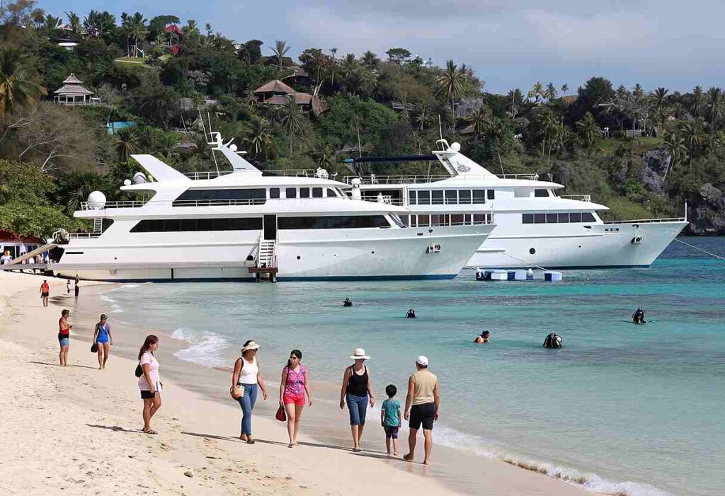 Two large white yachts docked near a sandy tropical beach while people stroll along the shoreline and clear turquoise water sparkles nearby.