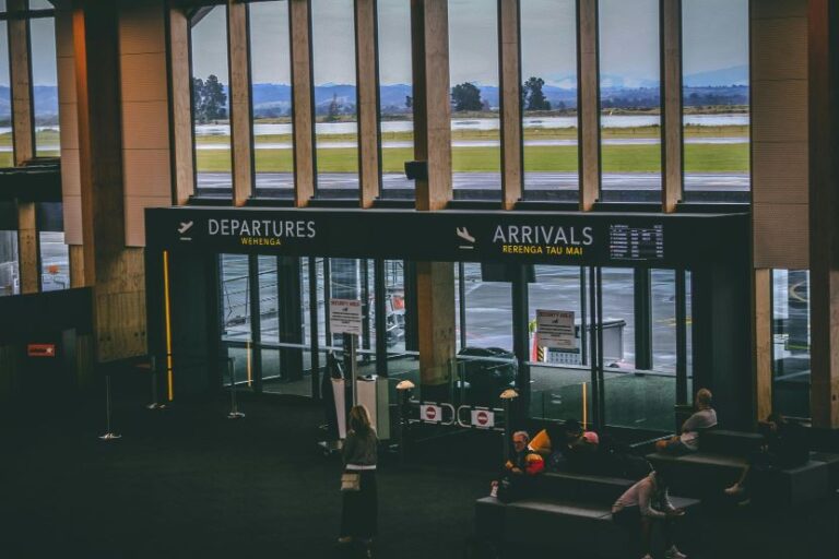 Airport terminal interior with departures and arrivals signage; people sit in couches while large windows overlook runways and hills beyond.