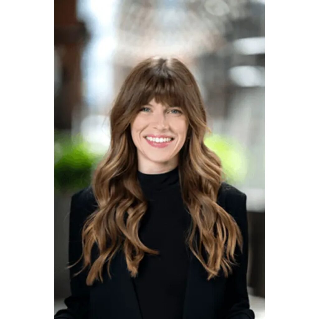 Smiling woman with long wavy brown hair wearing a black top and blazer, outdoors. (Professional headshot)