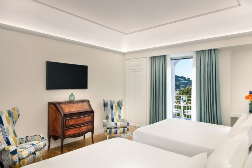 Bright hotel bedroom with two white beds, a wooden dresser, two colorful striped armchairs, and a balcony view through turquoise curtains.