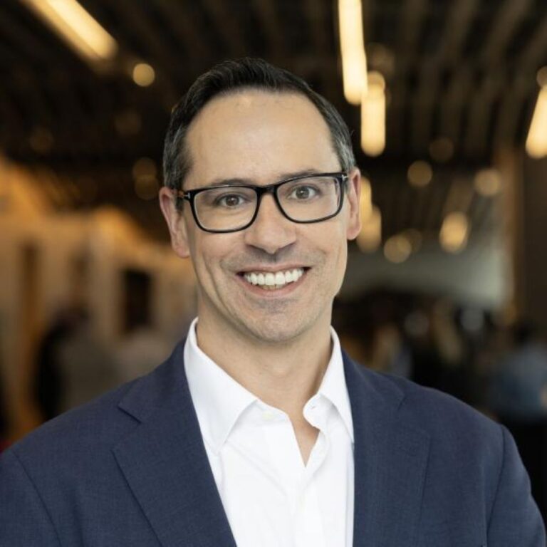 Professional portrait of a man wearing a navy blazer and white shirt with glasses, smiling at the camera in a conference-like setting.