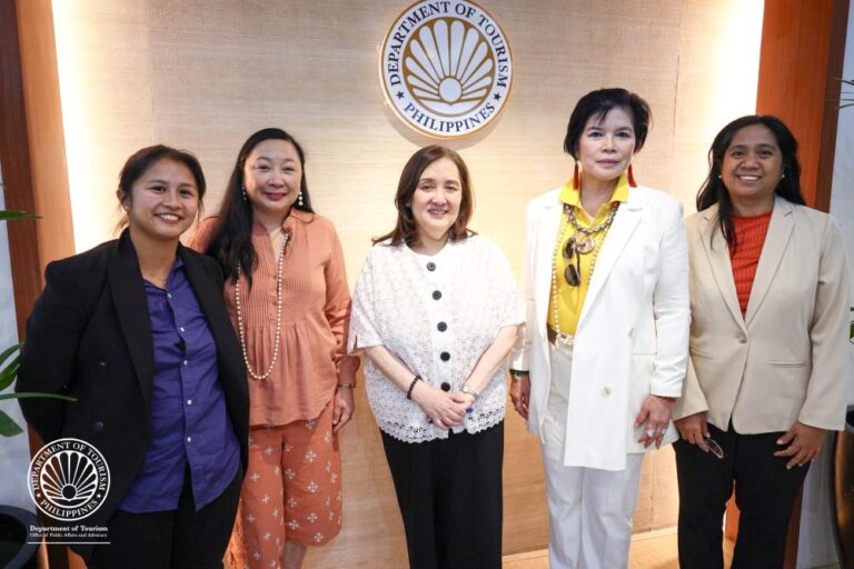 Five women in professional attire posing together in front of a Department of Tourism Philippines seal on a wood-paneled wall.